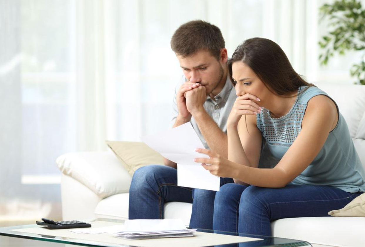 Couple sitting on couch with worried expressions, reviewing financial documents in a bright living room.