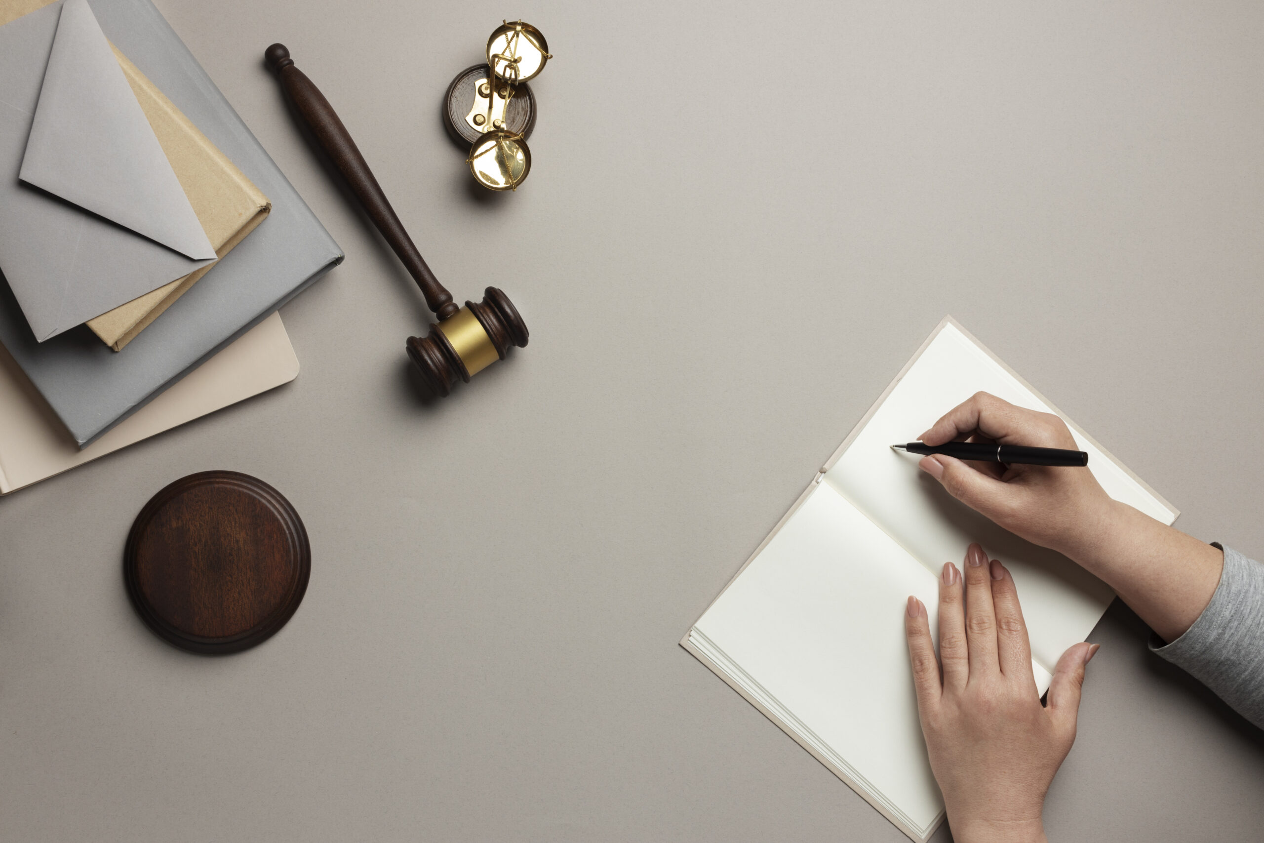 Overhead view of hands writing in notebook with gavel, envelopes, and scales symbolizing justice and legal work.