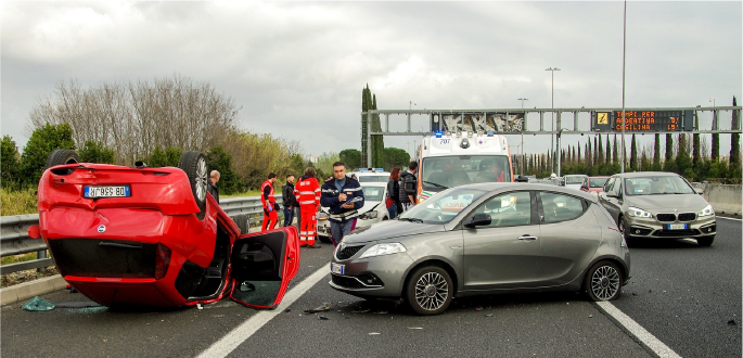 Traffic accident scene on a highway with flipped red car, emergency responders, and an ambulance on site.