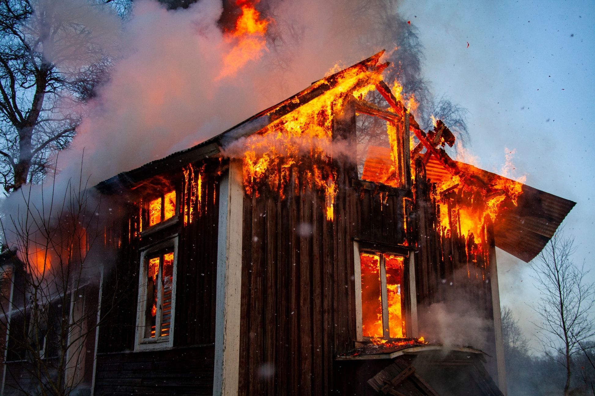 Burning wooden house engulfed in flames and smoke against a clear blue sky.