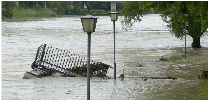 Flooded street with submerged fence and lampposts, in a natural setting with trees and water flow.