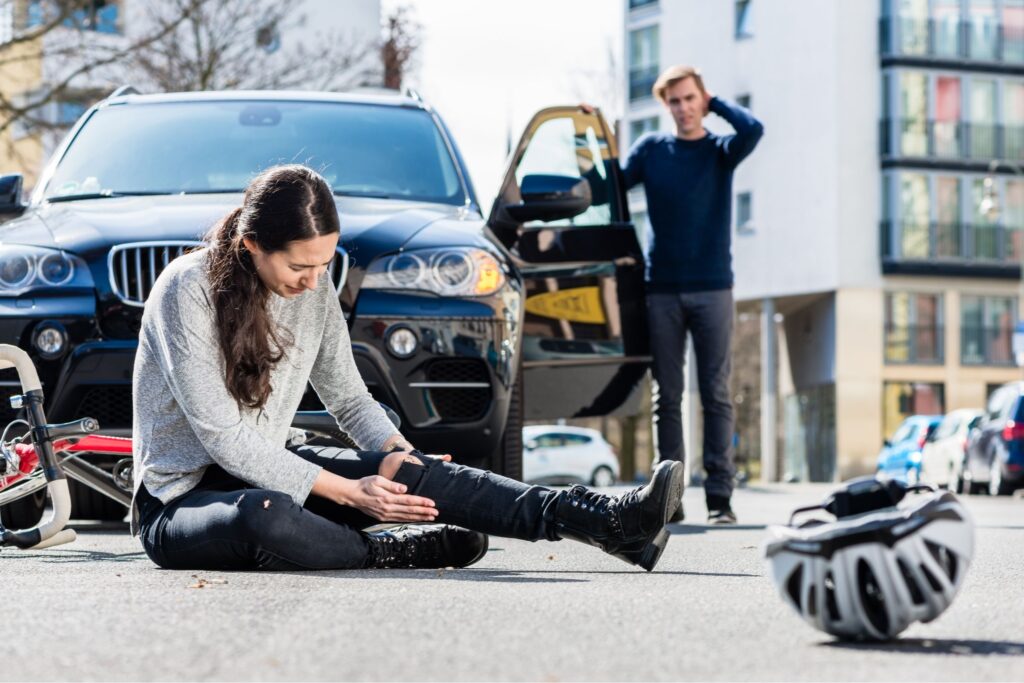 Woman injured in a bicycle accident with car, driver concerned. Helmet on ground, urban street setting.
