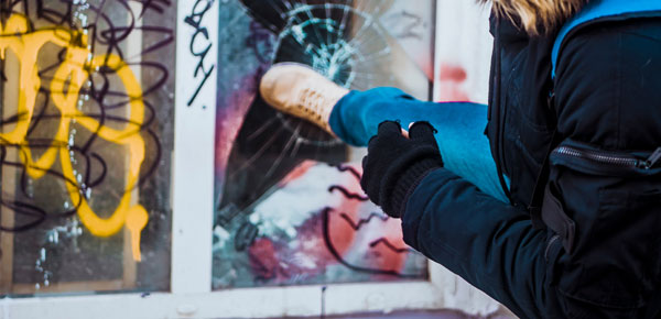 Person kicking through glass window with graffiti in background, wearing jeans and black jacket.
