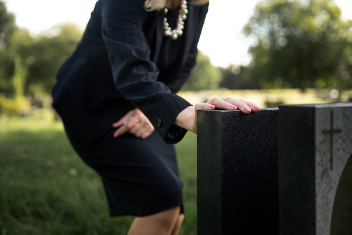 Person in black attire beside a gravestone, symbolizing reflection and remembrance in a serene cemetery setting.