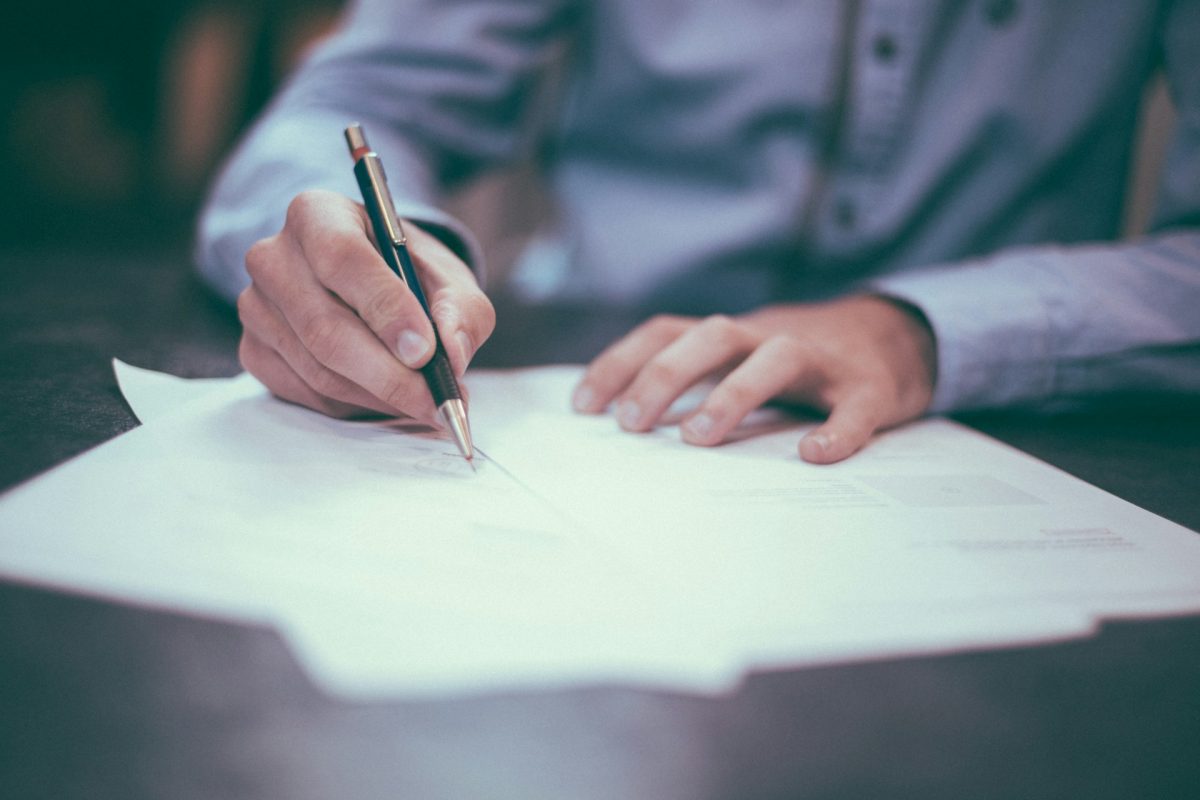 Person writing on paper at a desk, holding a pen, wearing a shirt, focused on work or study.