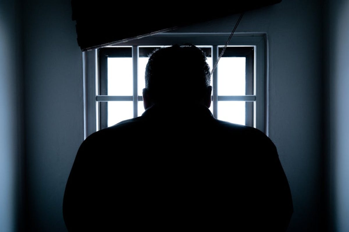 Shadowed figure gazing out a barred window, suggesting solitude or confinement.