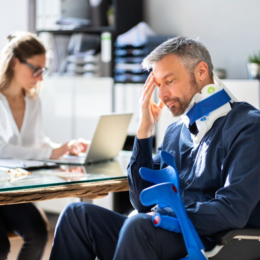 Man in wheelchair with neck brace looking stressed, while woman works on laptop at desk.