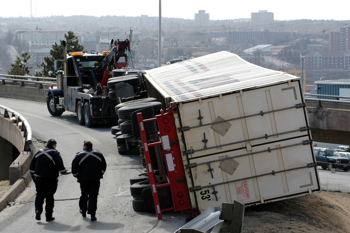 Overturned truck on highway ramp, tow truck and two people assisting, urban background.
