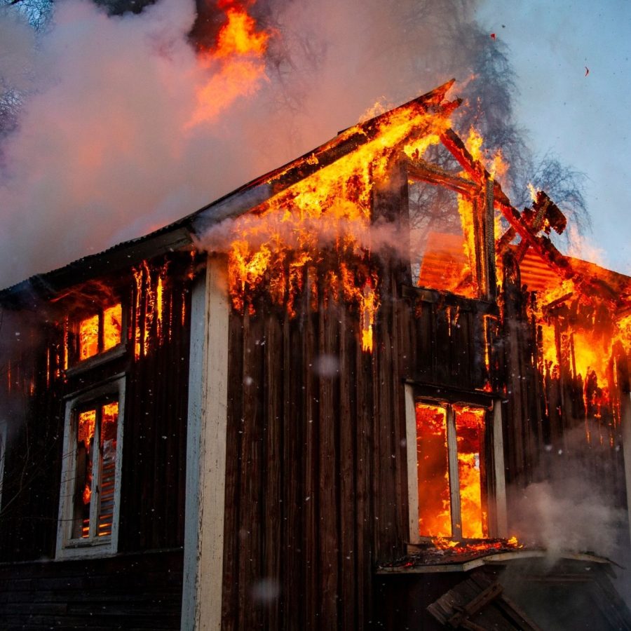 Burning wooden house engulfed in flames and smoke against a clear blue sky.