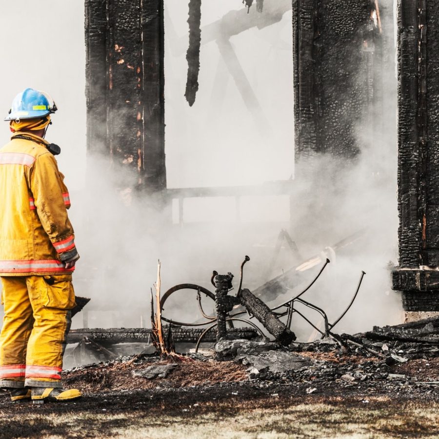 Firefighter in yellow suit assessing smoldering ruins of a burnt structure, surrounded by smoke.