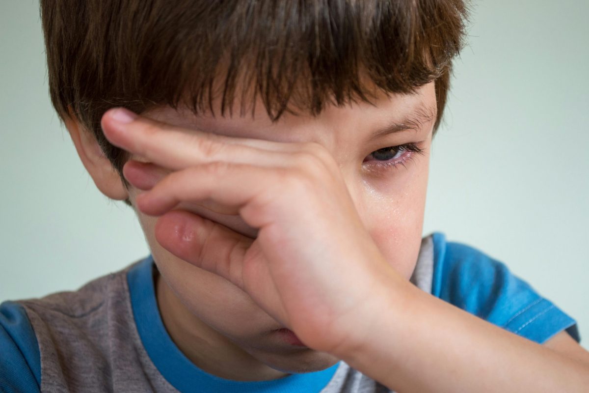 Child rubbing eyes, close-up of sad expression, wearing a blue and gray shirt.