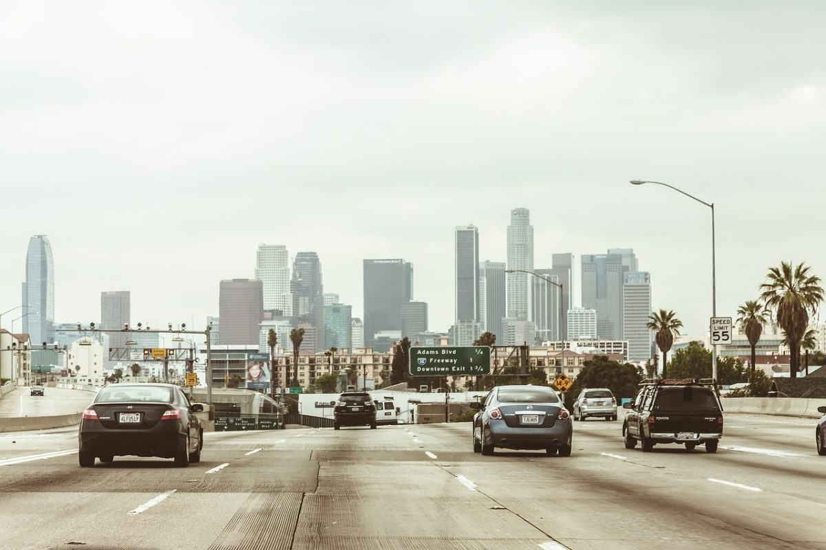 Cars driving towards downtown Los Angeles skyline under cloudy sky on freeway with palm trees and traffic signs.
