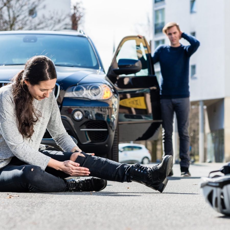 Woman injured on road after bike accident with car, while a concerned driver stands by.