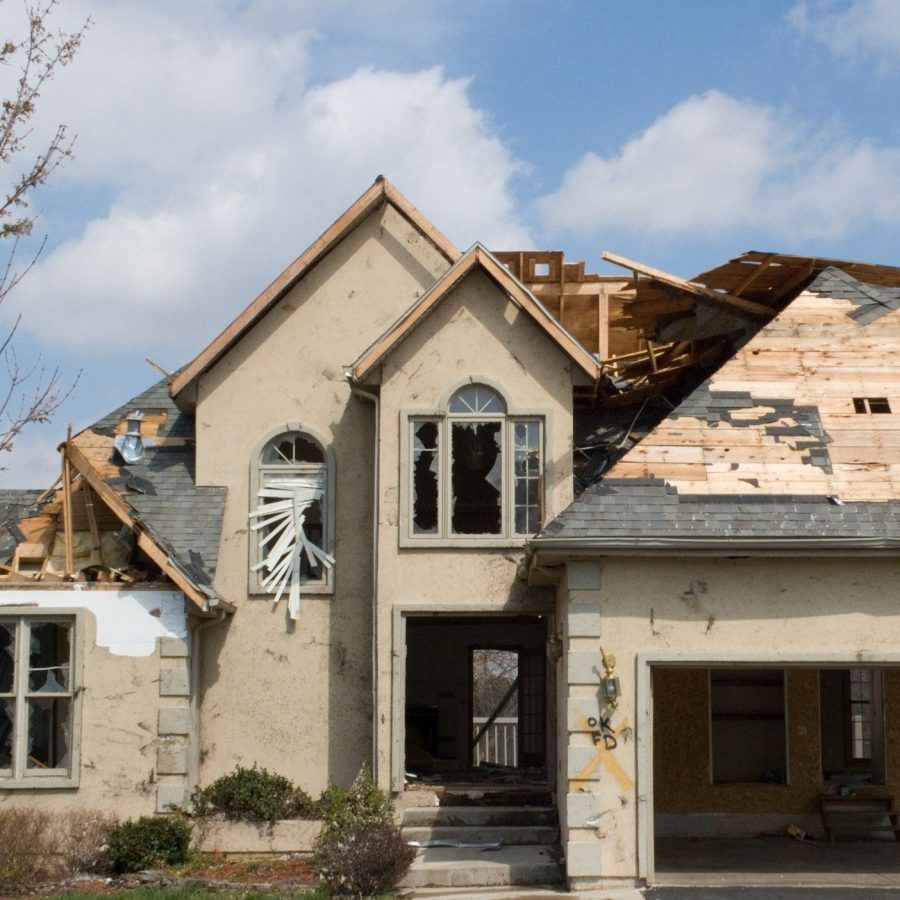 House with severe roof damage after storm, broken windows, and debris visible under a clear sky.