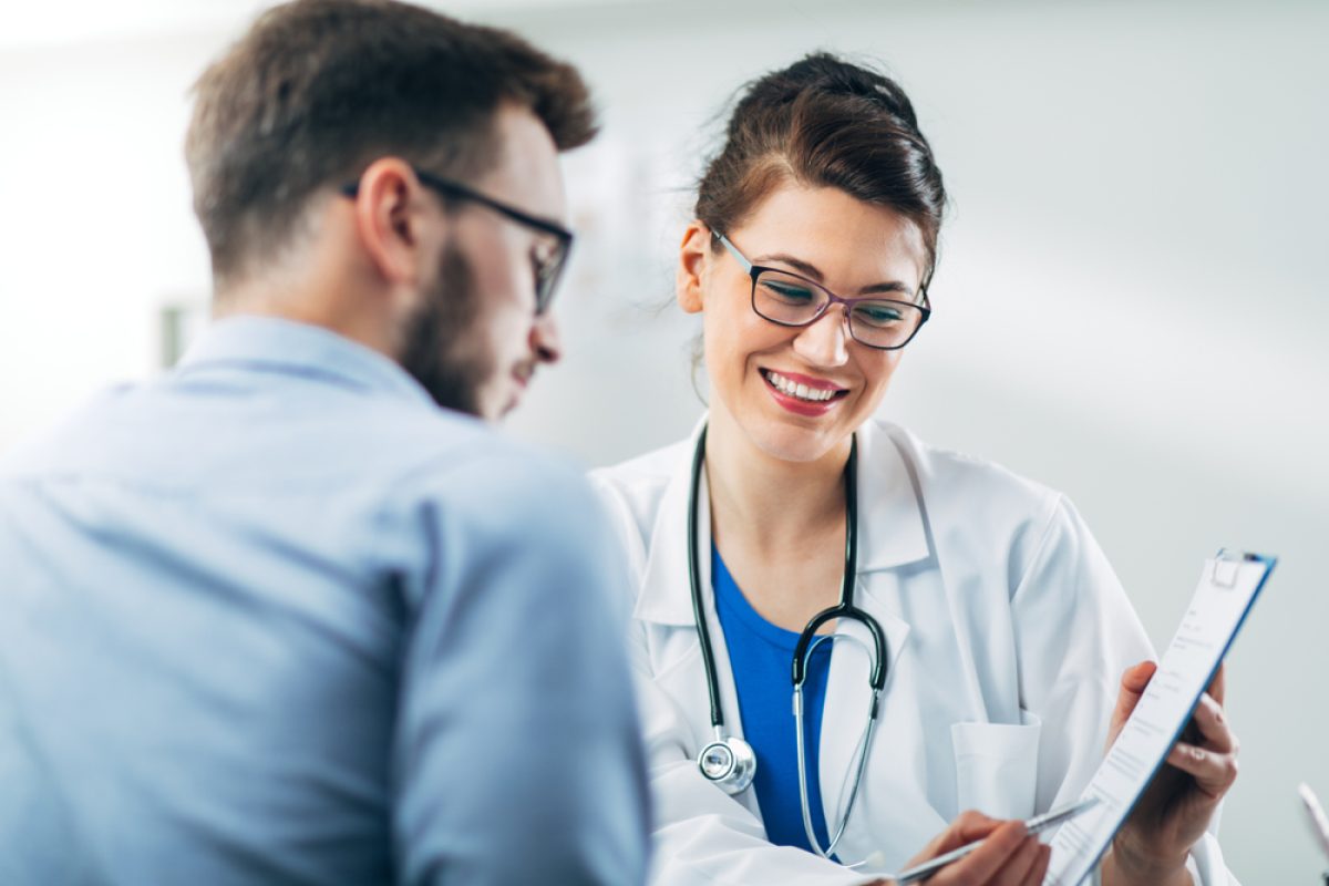Doctor consulting with a patient, smiling and showing medical results on a clipboard in a clinical setting.