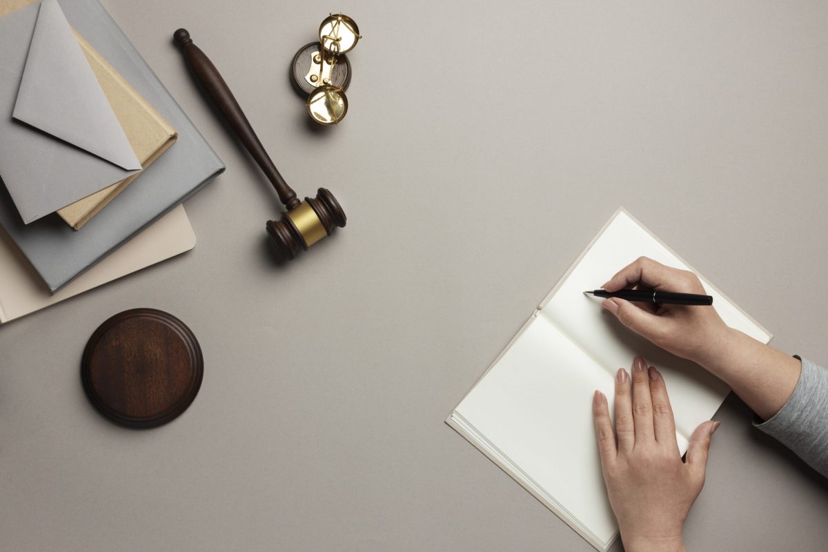 Overhead view of hands writing in notebook with gavel, envelopes, and scales symbolizing justice and legal work.