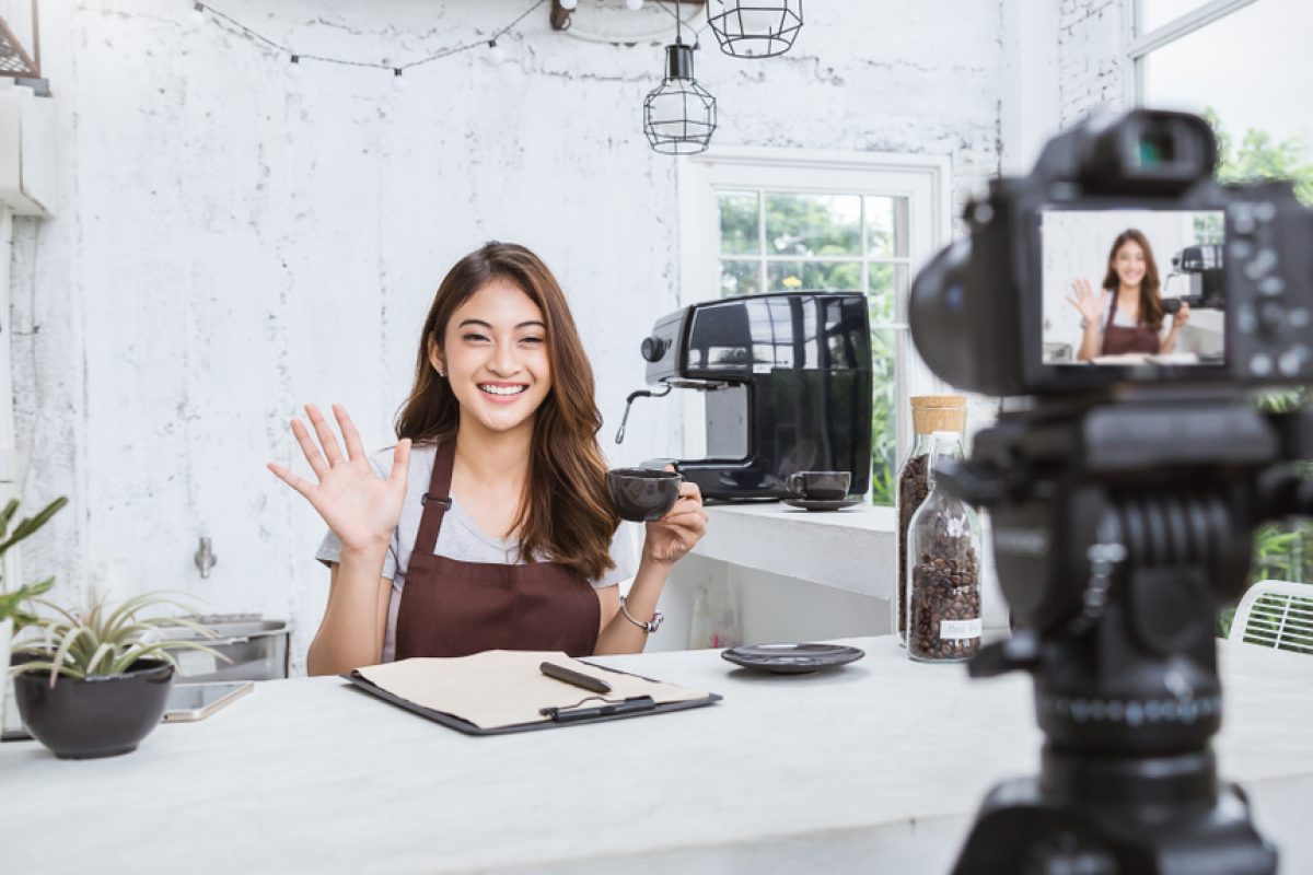 Woman in apron filming a coffee tutorial in bright kitchen, smiling and waving at camera.