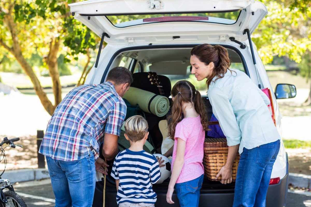 Family packing car for a road trip with picnic basket and camping gear on a sunny day.