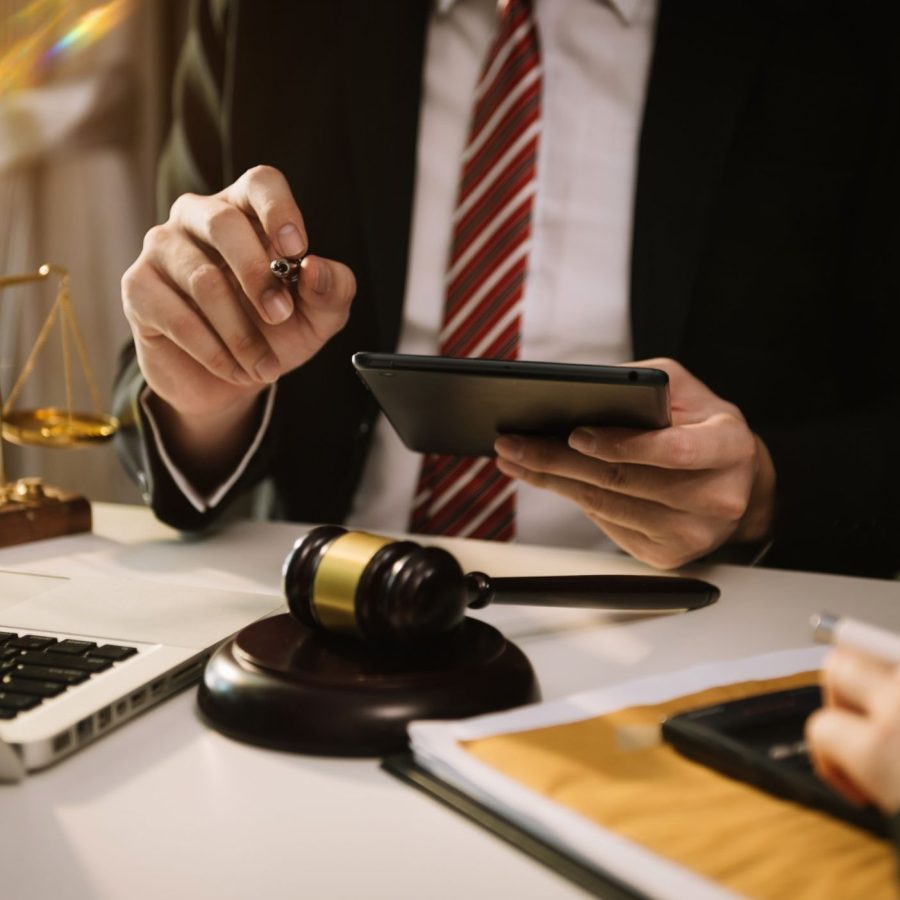 Lawyer in a suit using a tablet at a desk with scales, gavel, and laptop, representing legal consultation.