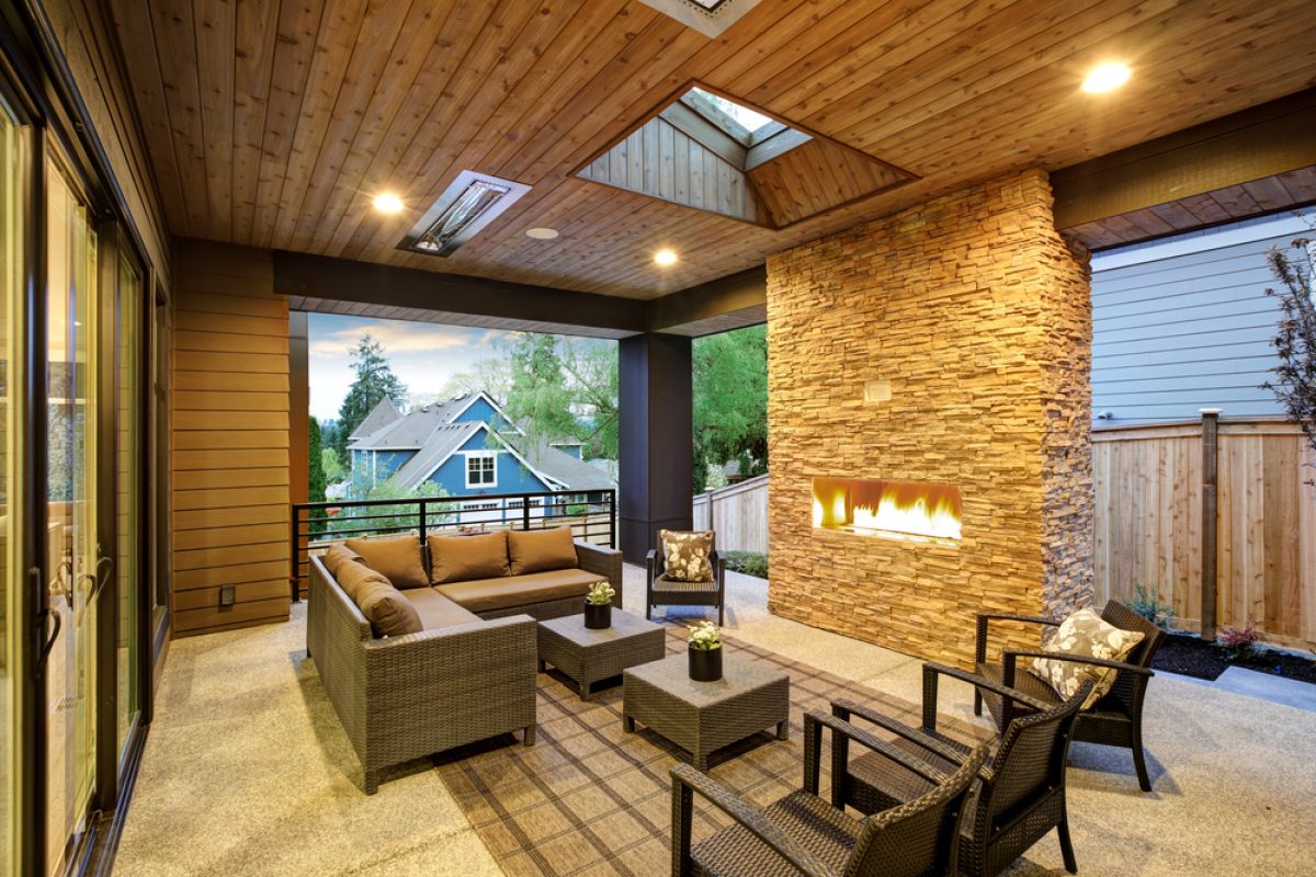 Modern covered patio with cozy wicker furniture, stone fireplace, and warm lighting, overlooking a suburban neighborhood.