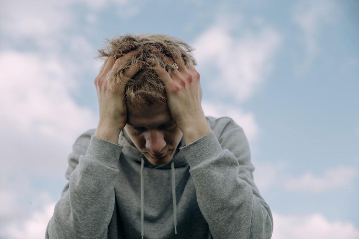 Person in gray hoodie holding head, looking stressed against a cloudy sky background.