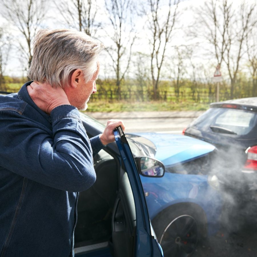 Man holding neck after car accident on a road with two damaged vehicles, showing collision and possible injury.