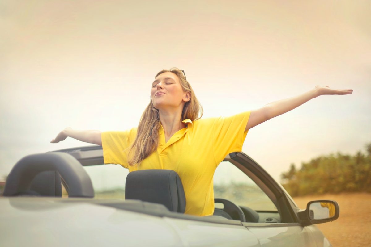 Woman joyfully stretching arms in a yellow shirt, standing in a convertible car, embracing freedom on a sunny day.