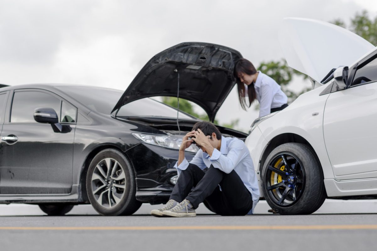 Two people in distress after a car accident, sitting near damaged vehicles with open hoods on the road.