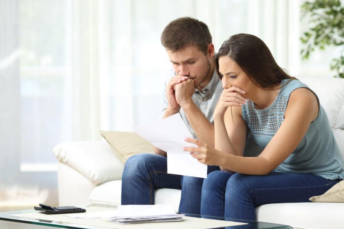 Couple sitting on couch with worried expressions, reviewing financial documents in a bright living room.